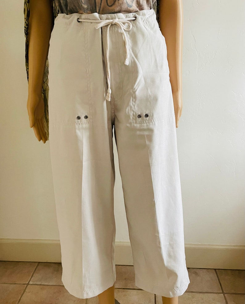 May include: White cropped pants with a drawstring waist and two pockets on the front. The pockets have two silver eyelets.