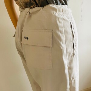 May include: A pair of light beige pants with a large patch pocket on the back. The pants have a drawstring waist and a zipper on the side seam.