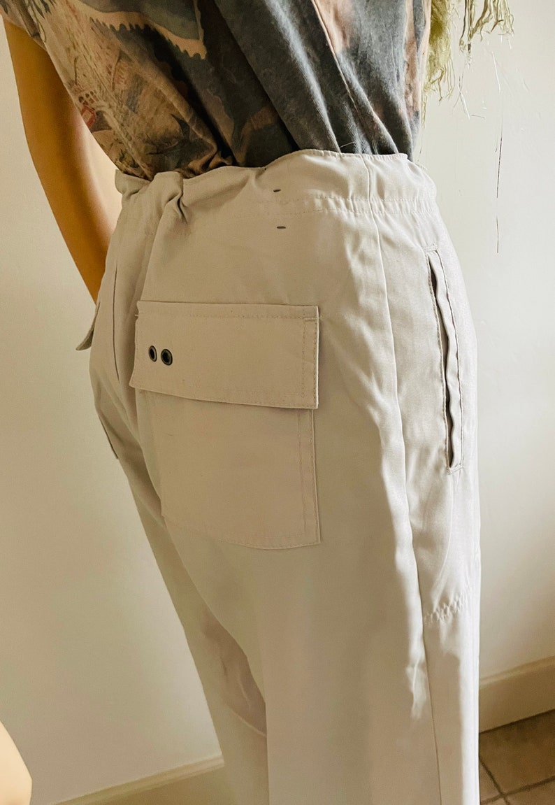 May include: A pair of light beige pants with a large patch pocket on the back. The pants have a drawstring waist and a zipper on the side seam.