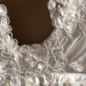 K&ouml;nnte beinhalten: Wei&szlig;er Satinstoff mit zarter Spitzenborte und Perlenbesatz. Die Spitze hat ein Blumenmuster.