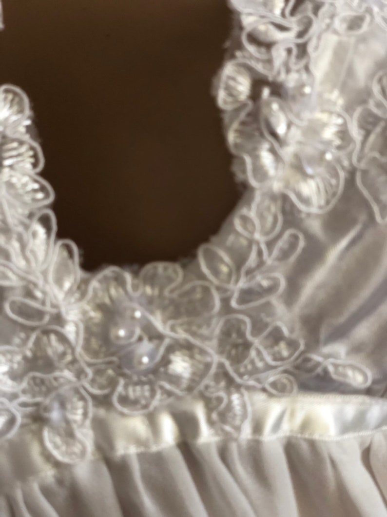 K&ouml;nnte beinhalten: Wei&szlig;er Satinstoff mit zarter Spitzenborte und Perlenbesatz. Die Spitze hat ein Blumenmuster.