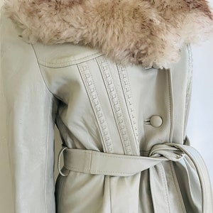 May include: A light grey leather coat with a large faux fur collar and a matching belt. The coat has decorative stitching on the front and a single button closure.