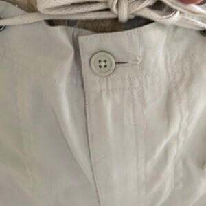 May include: Close-up of a white pair of pants with a button and a drawstring. The pants have a textured fabric and are made of a lightweight material.
