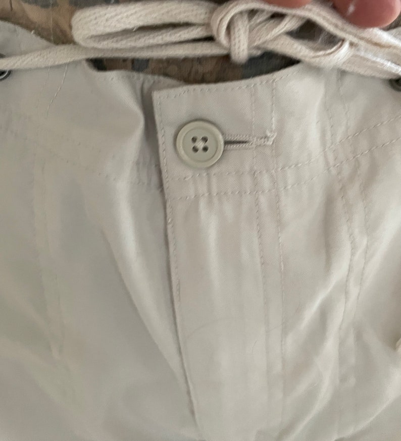 May include: Close-up of a white pair of pants with a button and a drawstring. The pants have a textured fabric and are made of a lightweight material.