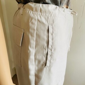 May include: A pair of light beige cargo pants with a single large pocket on the left side and a smaller pocket on the right side. The pants have a drawstring waist and a relaxed fit.