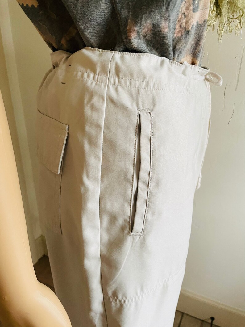 May include: A pair of light beige cargo pants with a single large pocket on the left side and a smaller pocket on the right side. The pants have a drawstring waist and a relaxed fit.