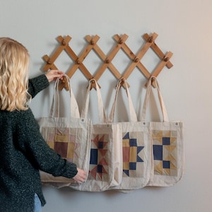May include: Four off-white canvas tote bags with geometric patchwork designs in shades of blue, yellow, and brown, hanging from a wooden accordion-style wall rack with multiple pegs against a plain light-colored wall.