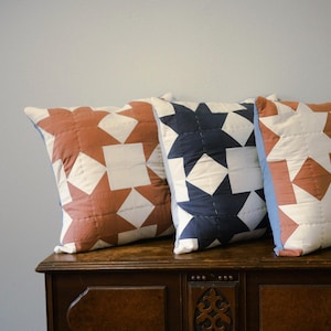 May include: Three square throw pillows featuring a quilted geometric star pattern. Two pillows display a terracotta and white color scheme, while the center pillow features a dark navy blue and white design. Each pillow has visible stitching.
