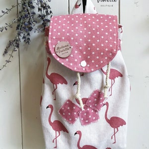 May include: A fabric backpack featuring a white base with a pink flamingo pattern. The flap is pink with white polka dots and a round wooden tag labelled Madame Violette. It includes a white drawstring closure and two frayed pink polka dot fabric accents.