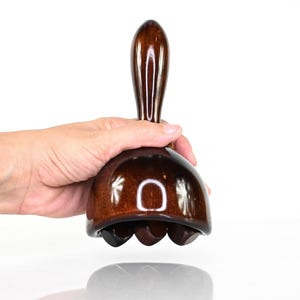 Könnte beinhalten: Ein dunkelbraunes, poliertes Handmassagegerät aus Holz mit einem glatten, abgerundeten Griff und einer kuppelförmigen Basis. Die Unterseite der Basis enthält drei kleine, kreisförmige Holzrollen, die zum Rollen über eine Oberfläche bestimmt sind.