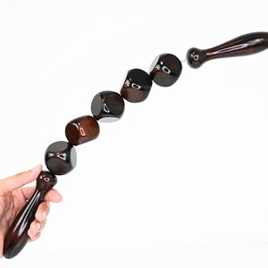 Könnte beinhalten: Ein dunkelbraunes Massagegerät aus Holz mit einem Griff und einer Reihe von verbundenen, abgerundeten, würfelförmigen Perlen. Die Perlen sind mit einem dünnen Metalldraht verbunden. Das Werkzeug befindet sich vor einem weißen Hintergrund.