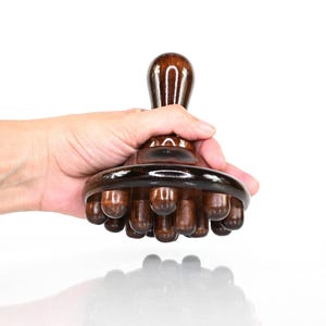 May include: A dark brown, polished wooden handheld massage tool featuring a rounded top handle and a circular base equipped with multiple smooth, rounded protrusions designed for applying pressure to the body during a massage session.