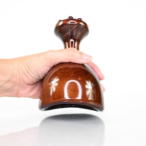 Può includere: Uno strumento per massaggi in legno lucido marrone scuro con una base liscia a forma di cupola e un manico ergonomico stretto sormontato da un piccolo pomello circolare. La superficie presenta una finitura lucida che riflette la luce.