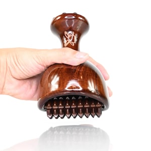 Può includere: Uno strumento per massaggi in legno lucido marrone scuro con un manico arrotondato e una base ampia a forma di cupola. La parte inferiore della base presenta una fila di piccoli denti in legno appuntiti progettati per applicare pressione.