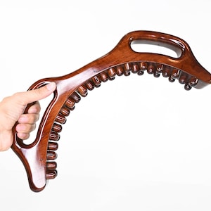 May include: A brown wooden back scratcher with a curved handle and rows of small wooden knobs. The scratcher is held in a hand.