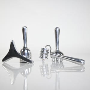 Può includere: Un assortimento di strumenti per massaggio color argento, tra cui una forma triangolare, due strumenti a forma di fungo e un piccolo rullo scanalato con un manico curvo. Gli strumenti sono disposti su una superficie riflettente.