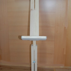 Puede incluir: Un caballete de madera con un acabado marrón claro. El caballete tiene una barra superior horizontal con dos soportes laterales ajustables y un poste central vertical con un panel deslizante. El caballete tiene una base con cuatro patas.