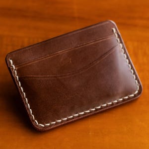以下が含まれることがあります： 茶色の革製カードケース。縁には白いステッチが施されています。カードケースには3つのカードスロットがあり、木製の背景に置かれています。革は滑らかで光沢のある仕上がりです。