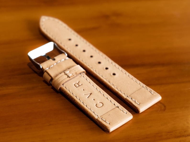May include: Two light brown leather watch straps with white stitching. The strap on the bottom has the text "C.A.R." embossed on it.