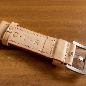 May include: A light brown leather watch strap with white stitching and a silver buckle. The strap is embossed with the initials "C.V.R."