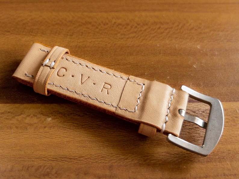 May include: A light brown leather watch strap with white stitching and a silver buckle. The strap is embossed with the initials "C.V.R."