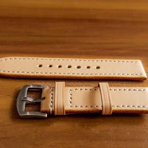 May include: A light brown leather watch strap with white stitching and a silver buckle.