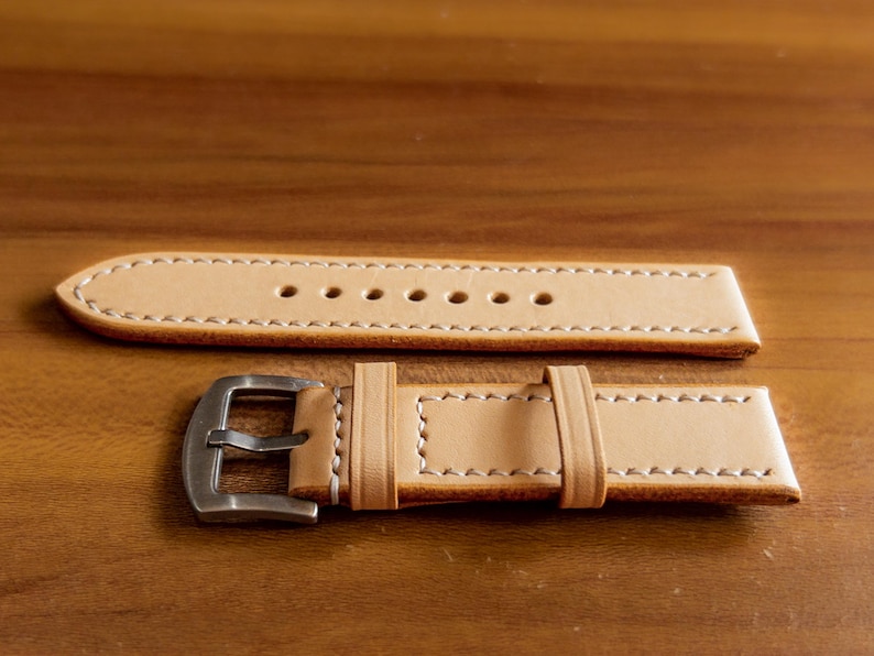 May include: A light brown leather watch strap with white stitching and a silver buckle.