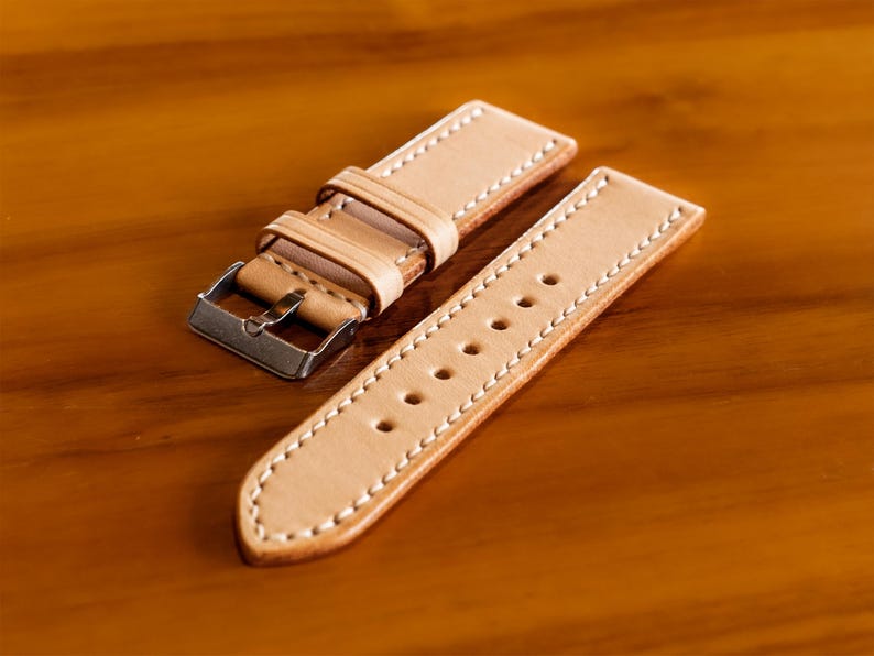 May include: A light brown leather watch strap with white stitching and a silver buckle.