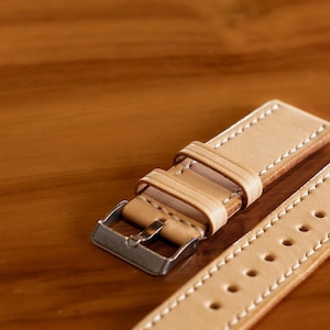 May include: A light brown leather watch strap with white stitching and a silver buckle.