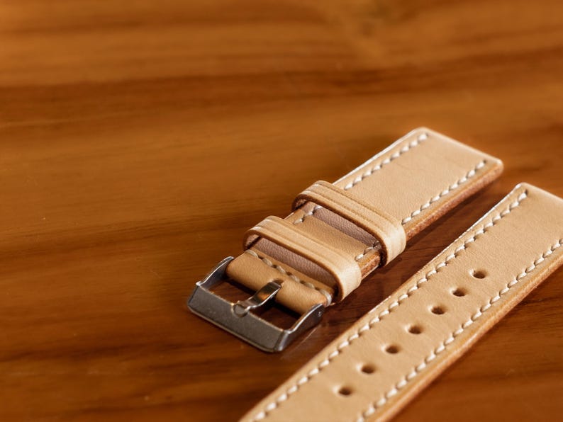 May include: A light brown leather watch strap with white stitching and a silver buckle.
