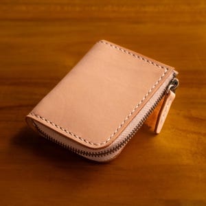以下が含まれることがあります： 明るい茶色の革製財布。長方形で、ジッパー開閉式。縁にはステッチが施されています。ジッパーは濃い金属色で、新品のように見えます。