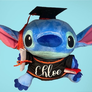 May include: Blue plush toy wearing a black graduation gown with an orange trim. The toy has a black graduation cap, a red tassel, and a diploma in its hand. The toy is holding a red book in its other hand. The gown has the name "Chloe" embroidered on the front.