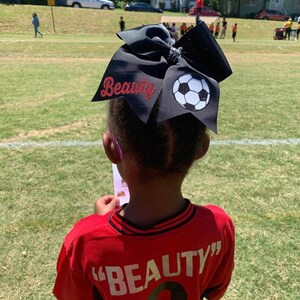 May include: A young person wearing a red soccer jersey with the word "BEAUTY" on the back. They are wearing a large black bow with a white soccer ball design and the word "Beauty" in red glitter on the bow.