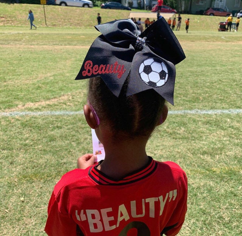 May include: A young person wearing a red soccer jersey with the word "BEAUTY" on the back. They are wearing a large black bow with a white soccer ball design and the word "Beauty" in red glitter on the bow.