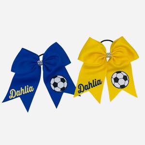 May include: Two cheer bows with a soccer ball design and the name "Dahlia" in gold glitter. The bows are blue and yellow.