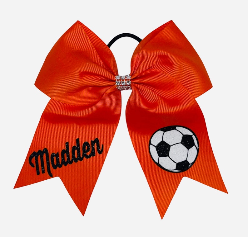 May include: Orange cheer bow with a black soccer ball and the name "Madden" in black glitter.