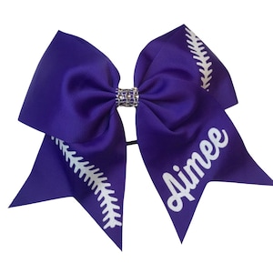 May include: Purple cheer bow with a white baseball stitch design and the name "Aimee" in white lettering. The bow has a rhinestone center.