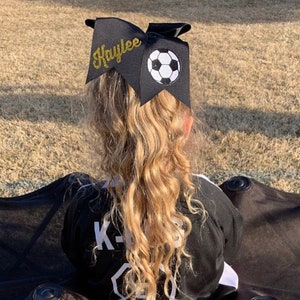 May include: Black hair bow with a gold glitter name "Kaylee" and a white and black soccer ball design.