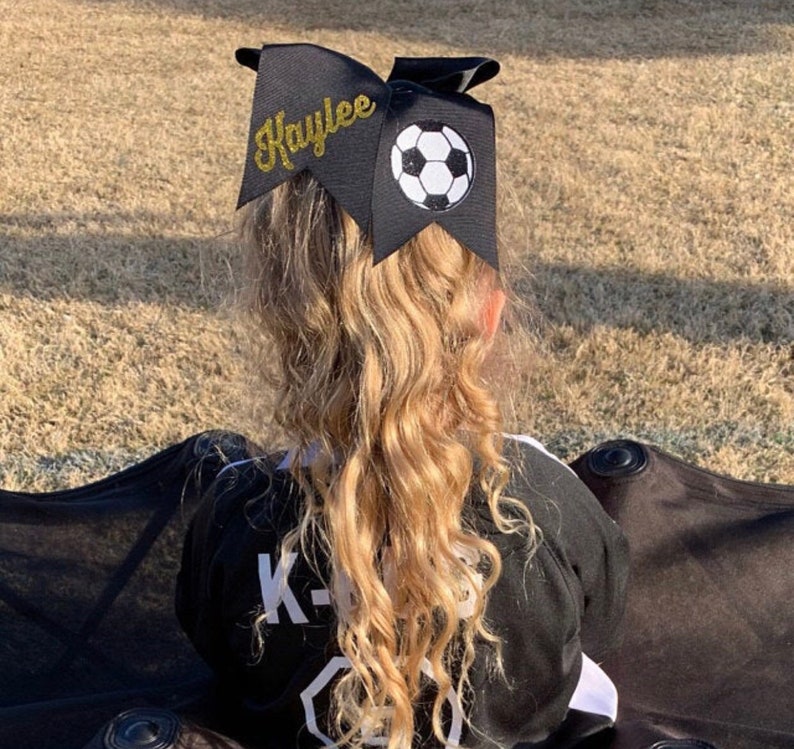 May include: Black hair bow with a gold glitter name "Kaylee" and a white and black soccer ball design.
