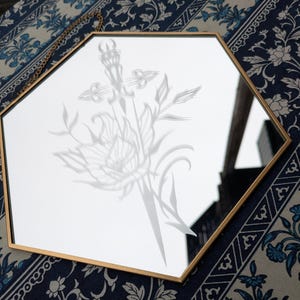 Może przedstawiać: Sześciokątne lustro ze złotą ramą. Lustro ma wytrawiony wzór miecza i elementów kwiatowych. Wisi na złotym łańcuszku. Tło to niebiesko-biała tkanina we wzory kwiatowe.