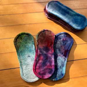 May include: A collection of reusable cloth menstrual pads in various colors, including green, red, and blue, with a tie-dye effect. The pads are stacked on a wooden surface, with three pads in the foreground and a stack of pads in the background.