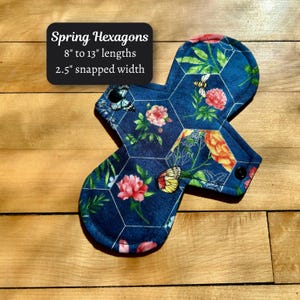May include: A reusable menstrual pad with a hexagonal pattern and a floral design featuring pink flowers, butterflies, and bees. The pad is dark blue and has black snaps. The text "Spring Hexagons" is displayed, with lengths from 8" to 13" and a 2.5" snapped width.