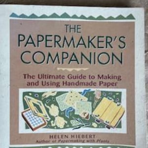 May include: A book titled "The Papermaker's Companion" with the subtitle "The Ultimate Guide to Making and Using Handmade Paper." The cover features a brown background with green and beige decorative elements and illustrations of papermaking tools.