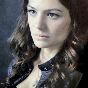 May include: A woman with long brown hair wearing a black leather jacket. She is looking off to the side with a serious expression.