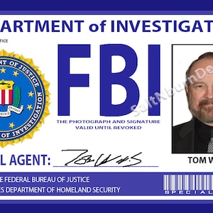 May include: A blue and white FBI identification card with the text "DEPARTMENT of INVESTIGATION" and "BUREAU OF JUSTICE" at the top. The card features a seal of the Federal Bureau of Investigation, the text "FBI" in large blue letters, and a photograph of a man with a beard. The card also includes the text "SPECIAL AGENT: Tom Willis" and a barcode with the text "SPECIAL AGENT".