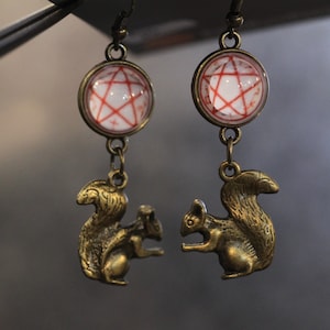 May include: A pair of earrings featuring a red pentagram design inside a glass dome, with a bronze squirrel charm dangling below each dome.