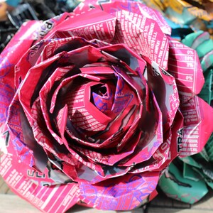 May include: A close-up of a handmade, large pink rose made from recycled aluminum cans. The petals are intricately folded, showcasing the printed labels and colors of the cans. Other colorful flowers are visible in the background.