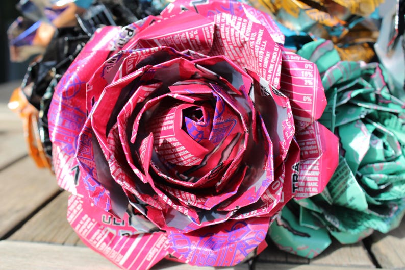 May include: A close-up of a handmade, large pink rose made from recycled aluminum cans. The petals are intricately folded, showcasing the printed labels and colors of the cans. Other colorful flowers are visible in the background.
