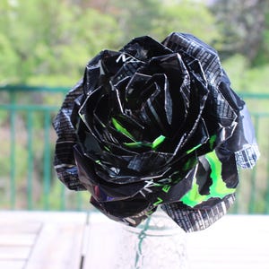 May include: A handmade black and green paper rose made from recycled materials. The flower has a layered petal design with green accents and is displayed in a clear glass vase. The background is a blurred outdoor scene.