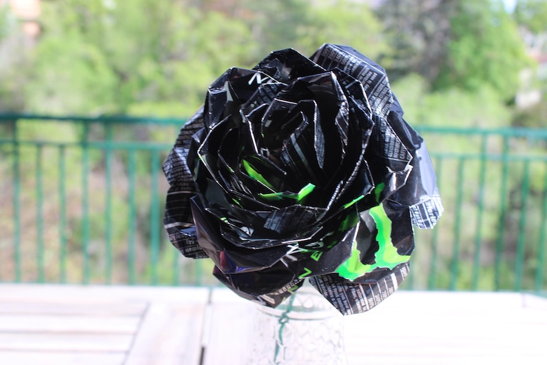 May include: A handmade black and green paper rose made from recycled materials. The flower has a layered petal design with green accents and is displayed in a clear glass vase. The background is a blurred outdoor scene.